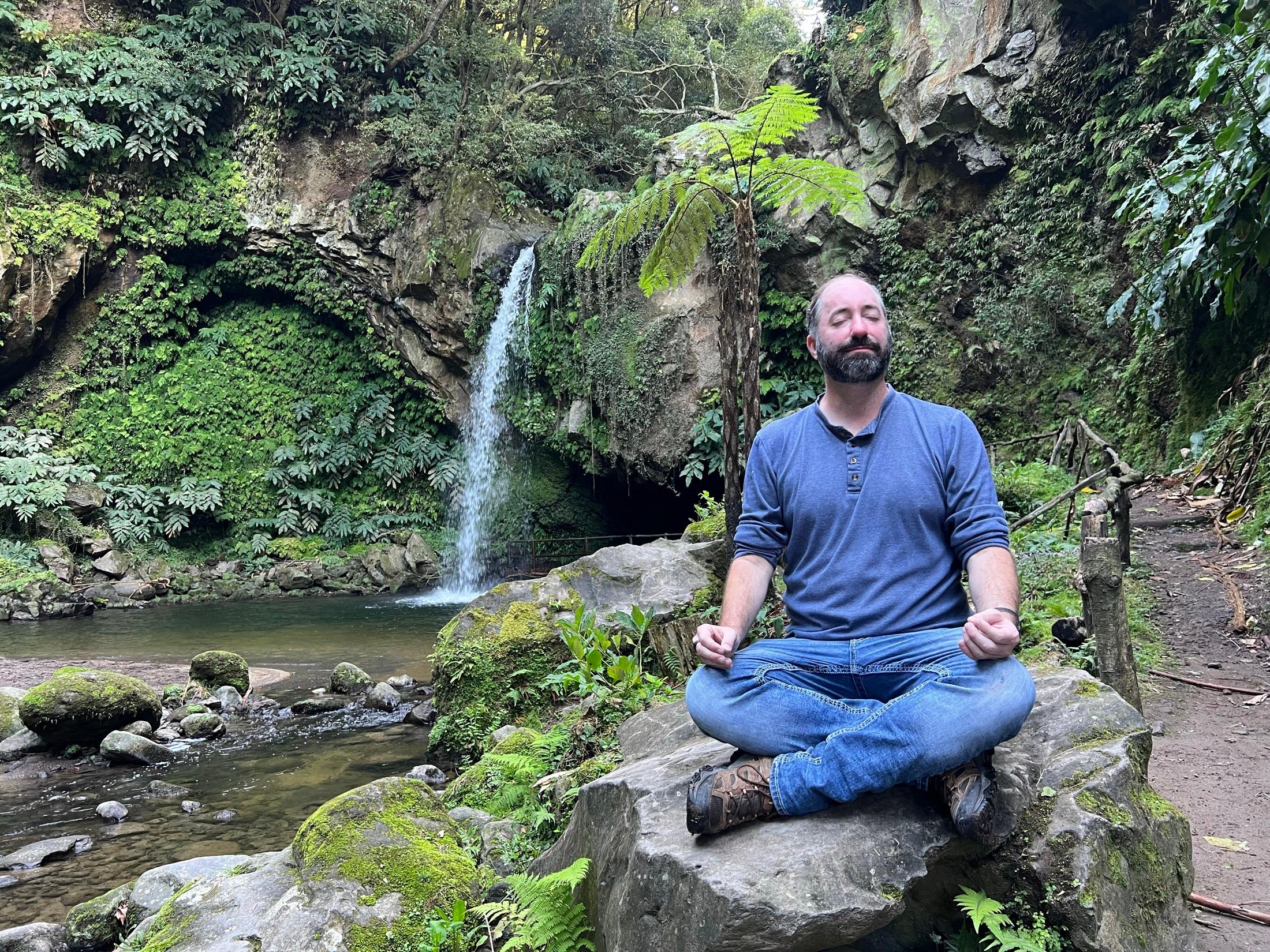 Trey Henderson meditating on a rock beside a waterfall in a green forest grotto